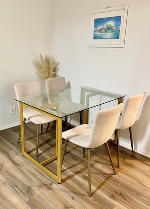 Un área de comedor con una mesa de vidrio y cuatro sillas, un jarrón con plumas de pampa y un cuadro en la pared.