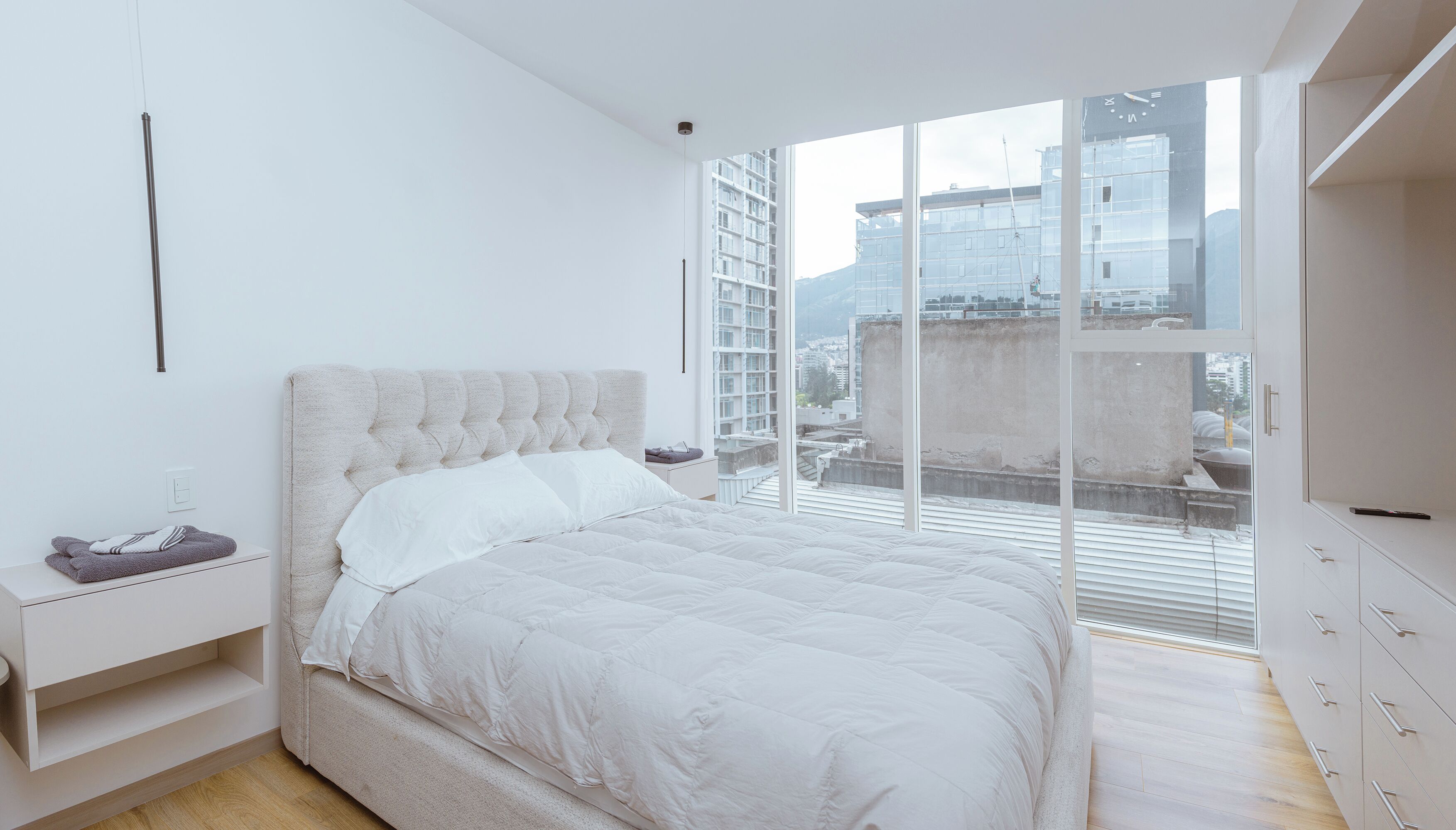 Un dormitorio moderno con una cama grande, una mesita de noche y vistas a la ciudad a través de ventanales del suelo al techo.