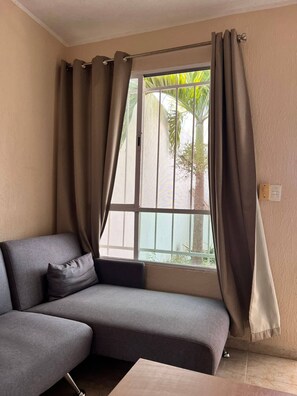 A corner room with a grey sofa, a window with curtains, and a view of greenery outside. - Nice House With Air Conditioning And Wifi (Mérida)