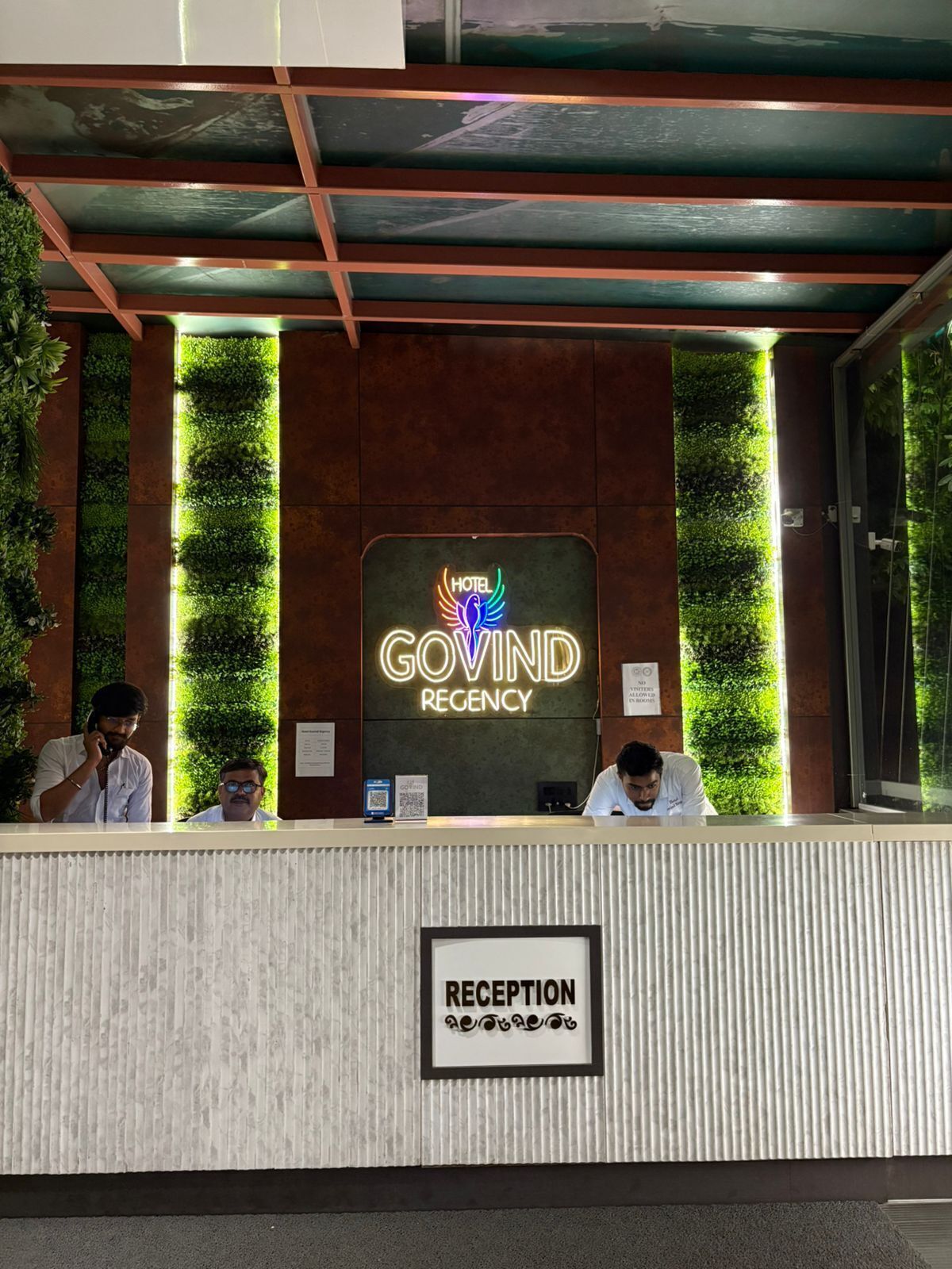 Mostrador de recepción de hotel con un letrero de neón para Govind Regency, un área de recepción y vegetación vertical.