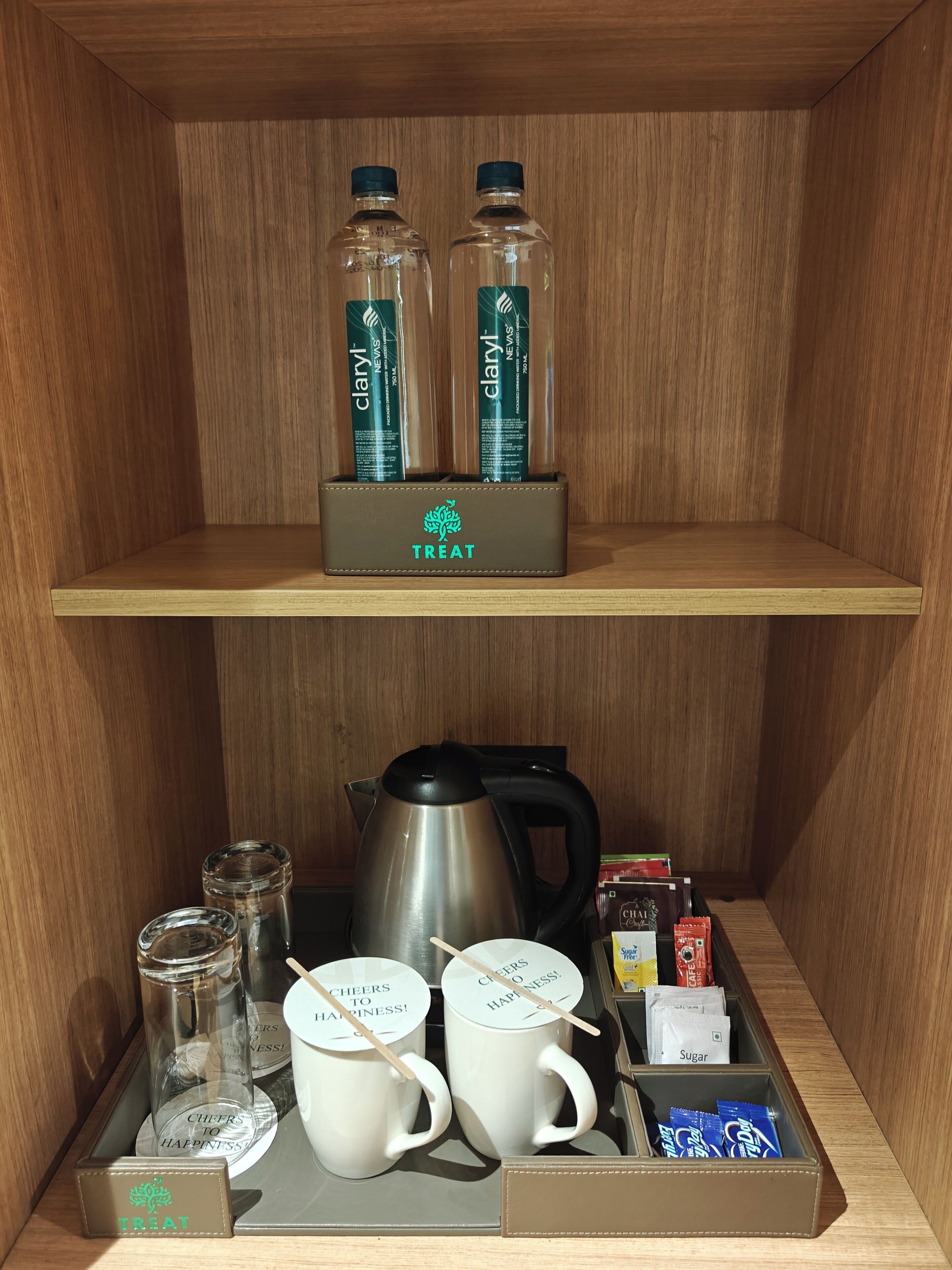 Une étagère en bois contenant des articles de toilette, notamment deux flacons de Claryl Nevis, une bouilloire, deux tasses blanches, un bocal en verre et une boîte étiquetée « Friandises ».