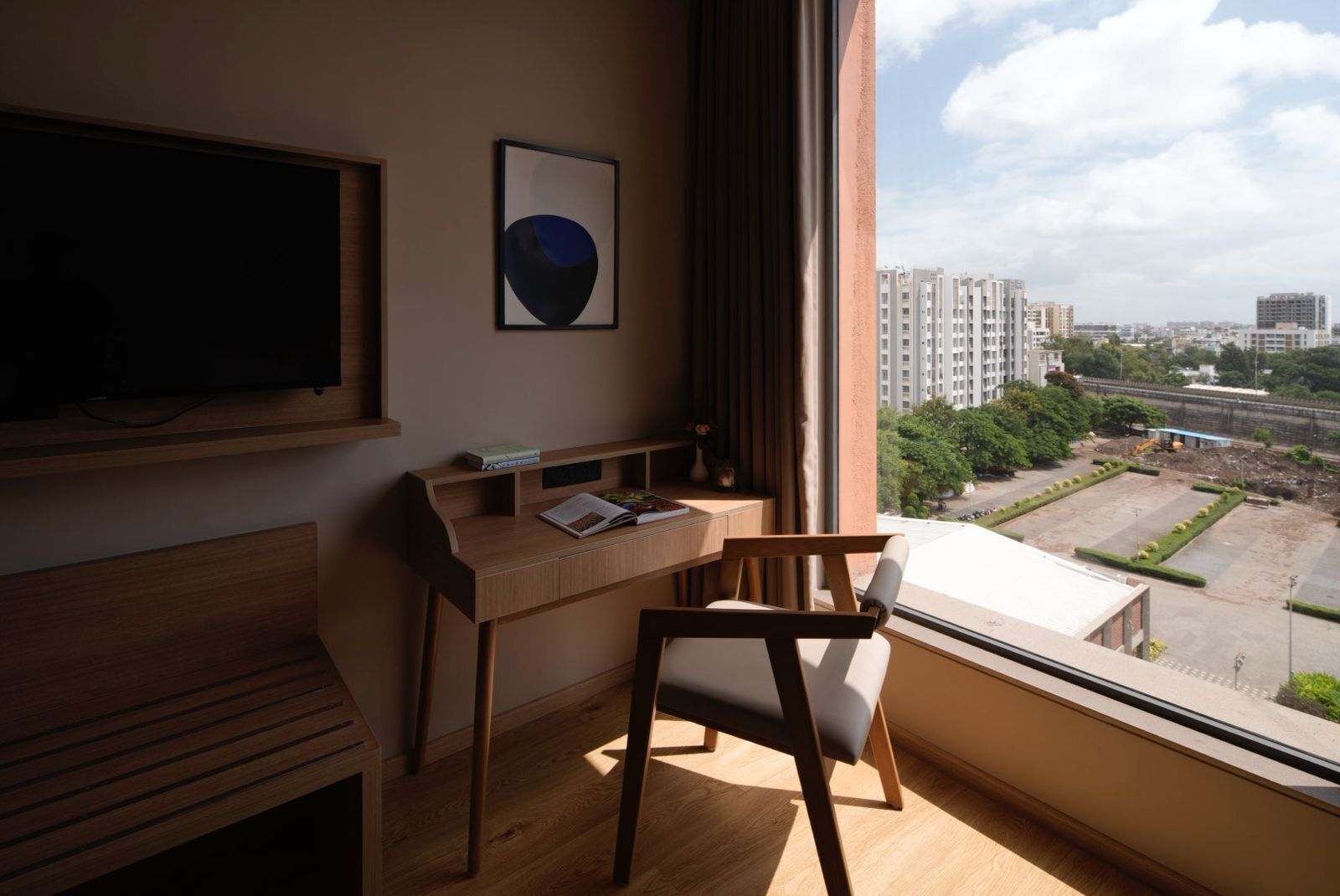 A modern room with a wooden desk, a chair, a flat-screen TV, and a large window offering a city view.