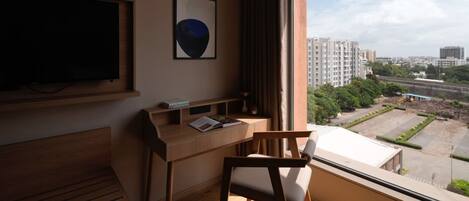 A modern room with a wooden desk, a chair, a flat-screen TV, and a large window offering a city view.