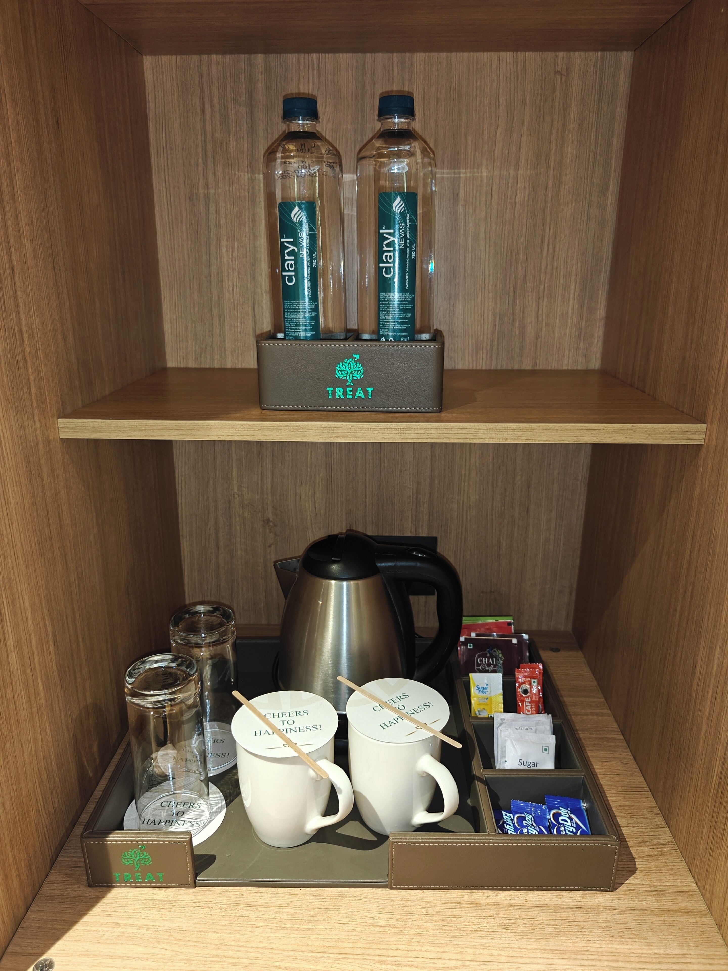 A hotel minibar with toiletries, including two bottles of Clary Perry Eau de Parfum, a coffee maker, sugar packets, and two mugs with "Cheers to Happiness!" labels.