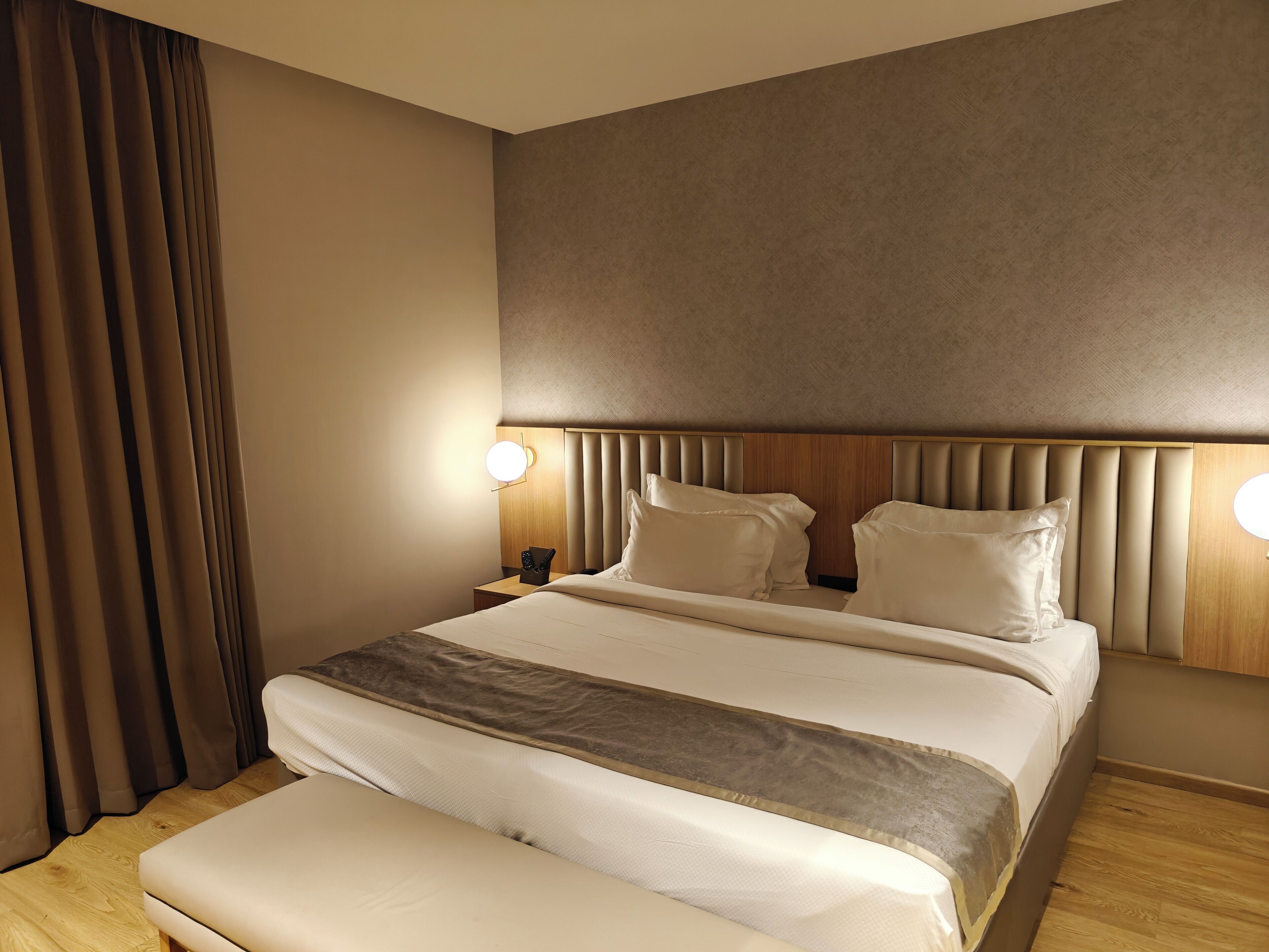 A modern hotel room with a large bed, two bedside tables, a nightstand, and a headboard with integrated lighting.