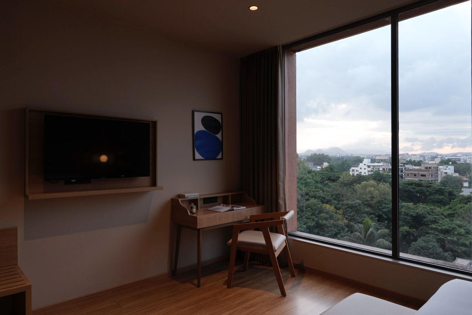 A modern room with a large window, a flat-screen TV mounted on the wall, a wooden desk, and a chair.