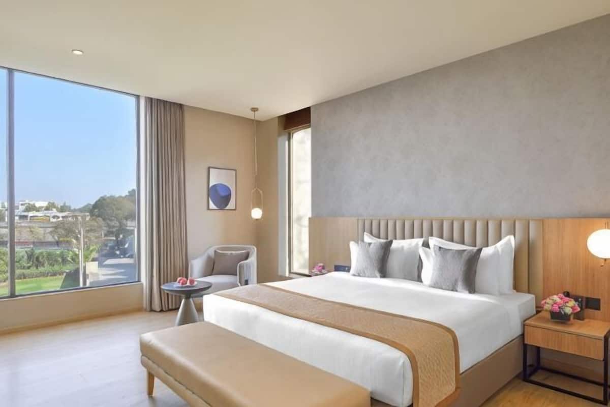 Habitación de hotel con una cama grande, una mesita de noche con lámpara, una silla, una pequeña mesa y vistas al exterior.