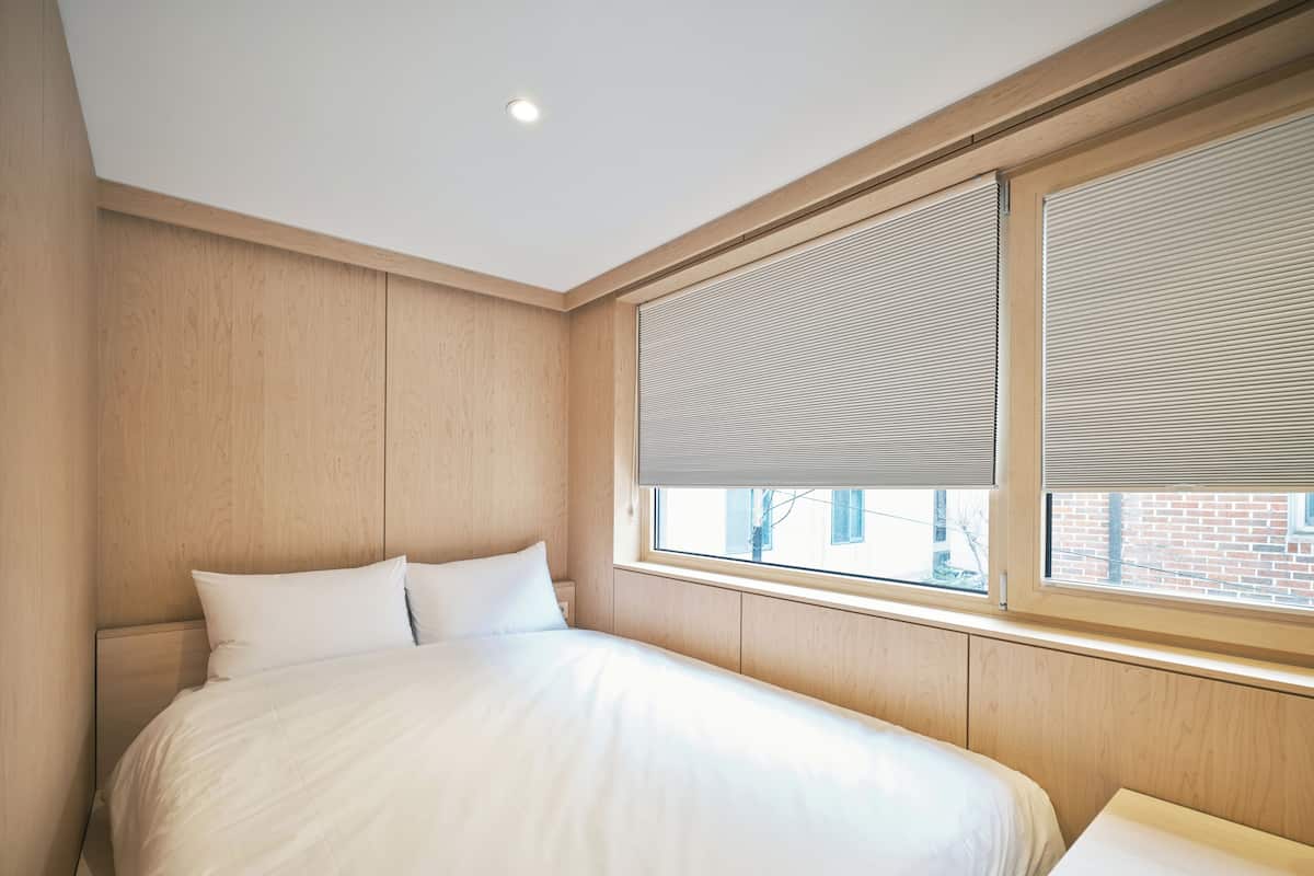 Un dormitorio con cama, paneles de madera, una ventana con persianas y una mesita de noche.