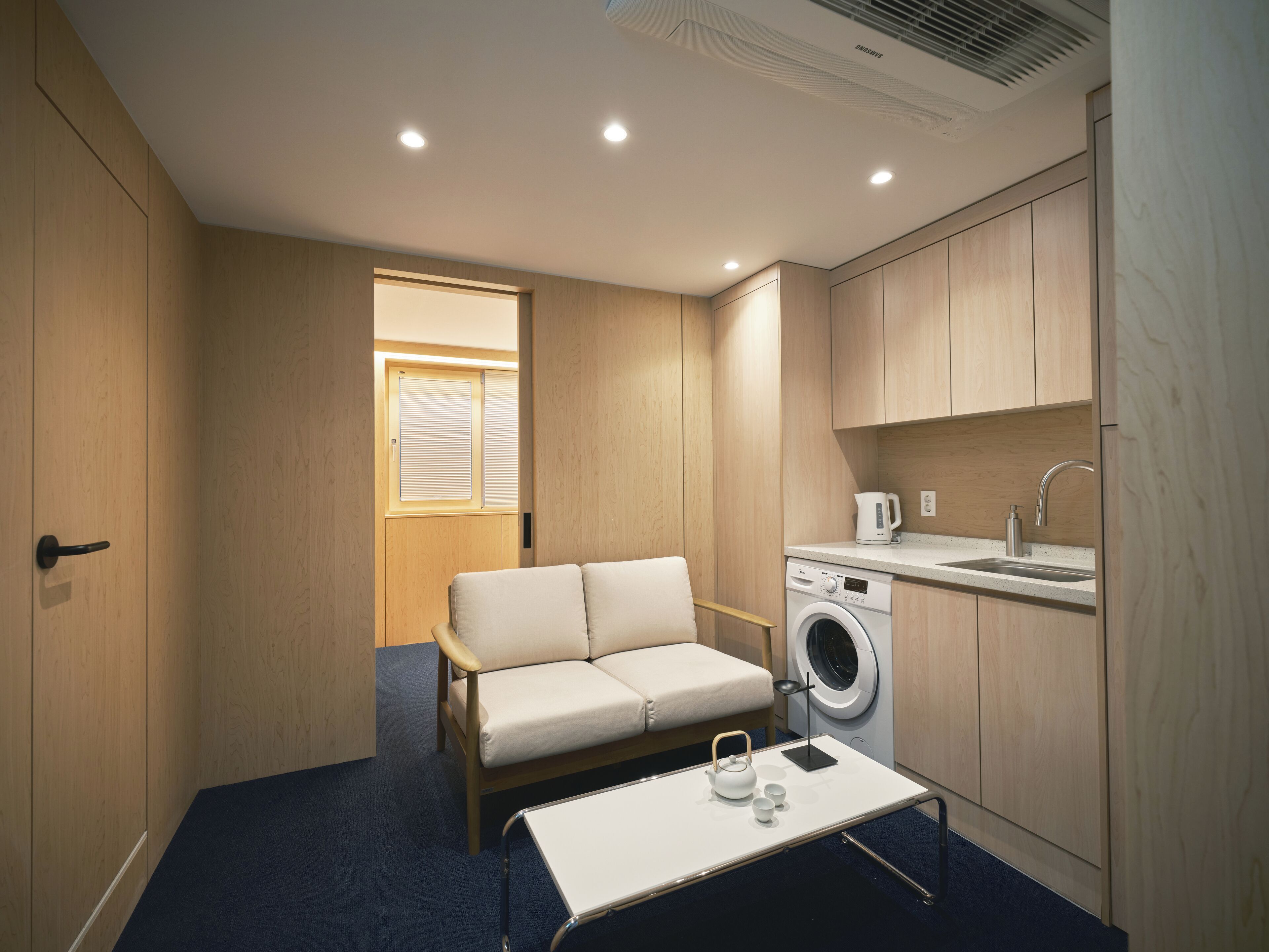 Habitación compacta con sofá, lavadora, lavamanos y ventana con persianas.