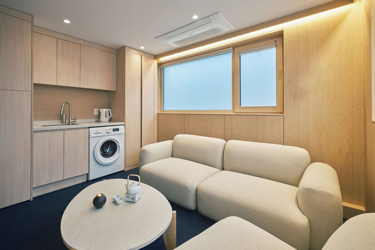 Un espacio habitable compacto con kitchenette, lavadora, sofá y una pequeña mesa redonda con tetera y tazas.