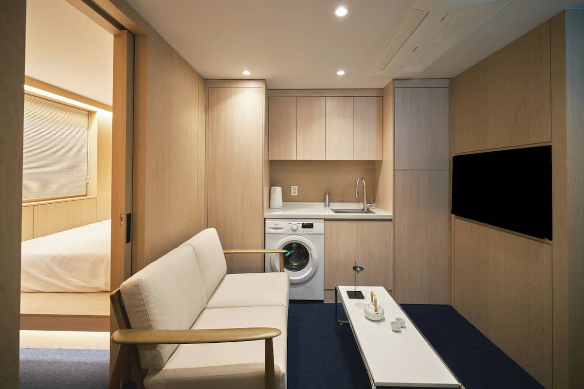 Habitación de hotel pequeña con sofá, lavadora, lavabo y televisor de pantalla plana montado en la pared.