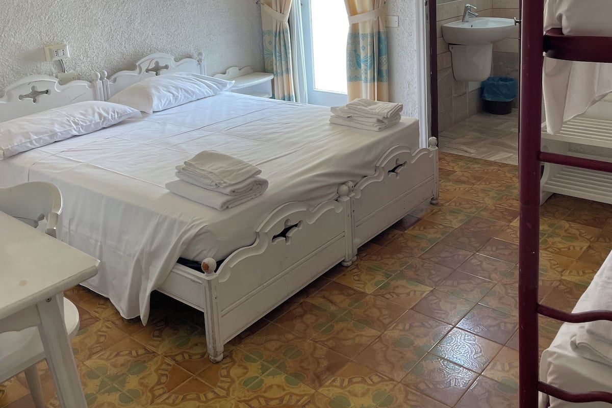Ein ordentlich eingerichtetes Schlafzimmer mit einem Bett, einem Nachttisch, einem Sessel, einem Schlafplatz und einem durchgehenden Türspalt sichtbaren Badezimmer.