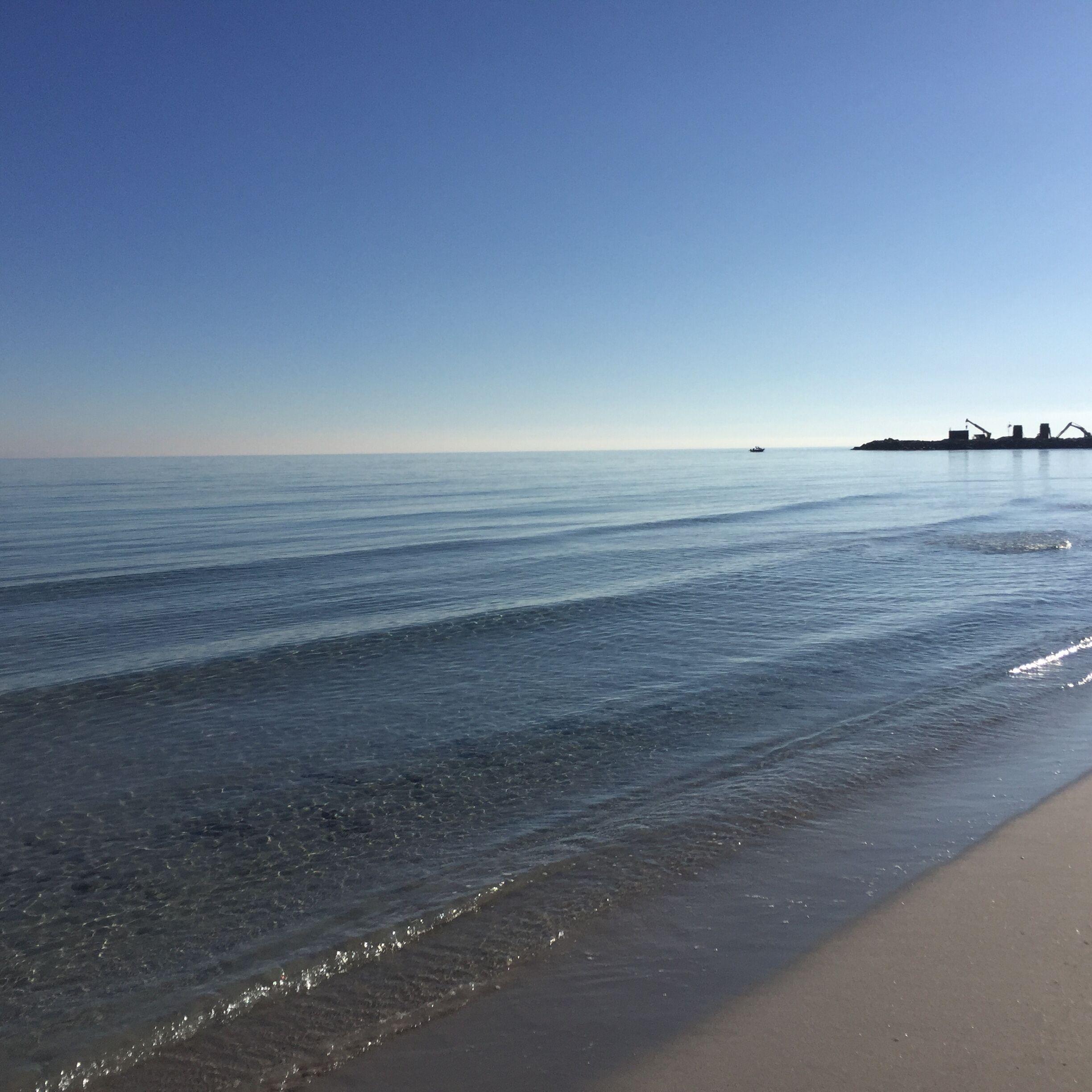 Ein Strand mit ruhigem Wasser und einer fernen Küstenlinie mit Industriegebäuden.