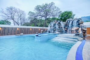 A swimming pool with a waterfall feature, surrounded by trees and a wooden wall.