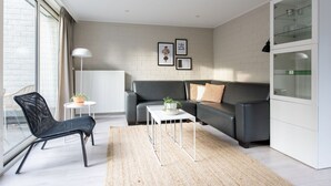 A modern living room with a grey sofa, a black chair, a small white table, and a potted plant. There are framed pictures on the wall and a radiator below. - Bungalow by Veluwe Sands (Kootwijk)