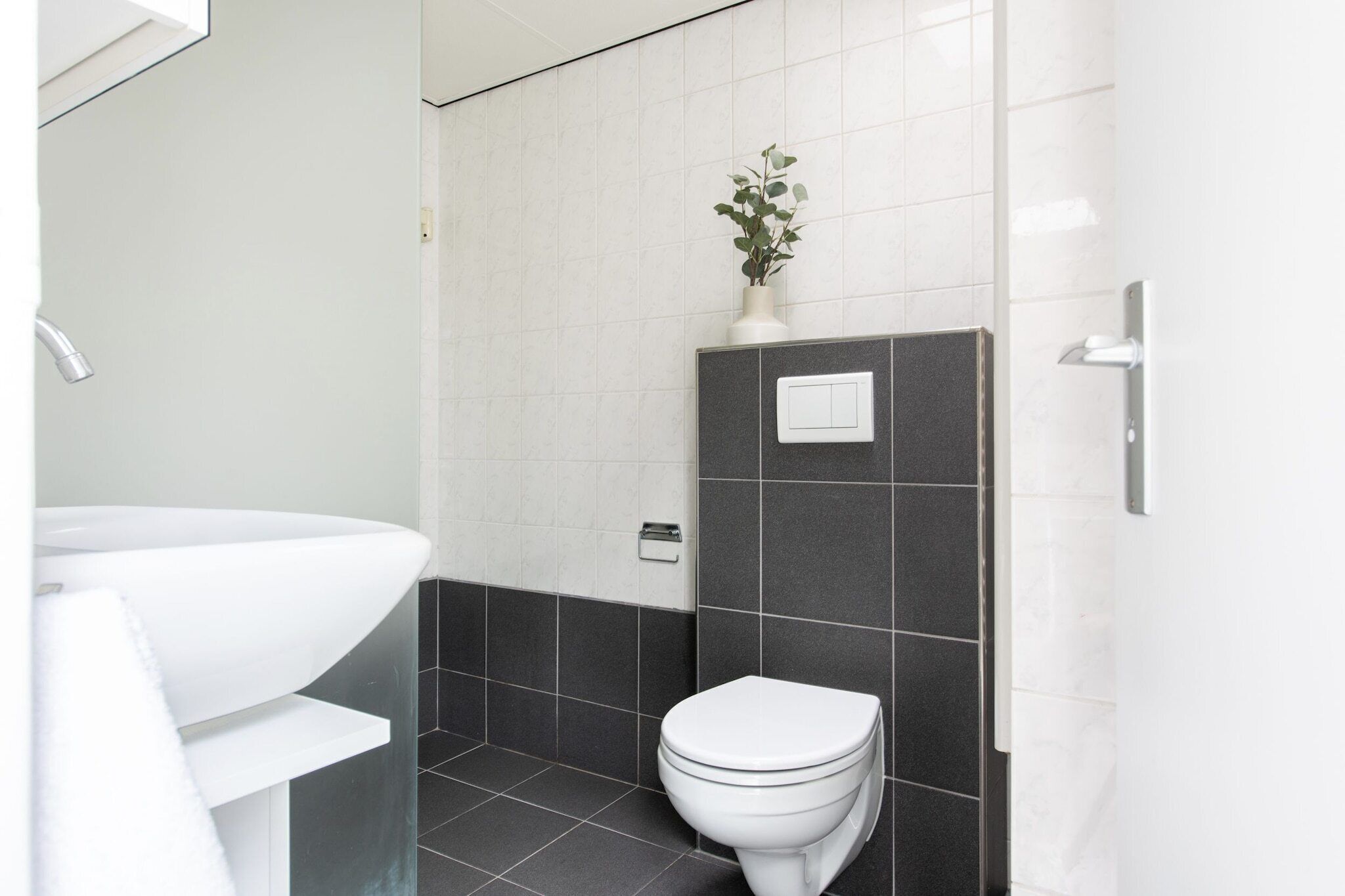 Une salle de bain moderne avec un lavabo blanc, des murs recouverts de carreaux foncés, des toilettes blanches et une plante en pot accrochée au mur.