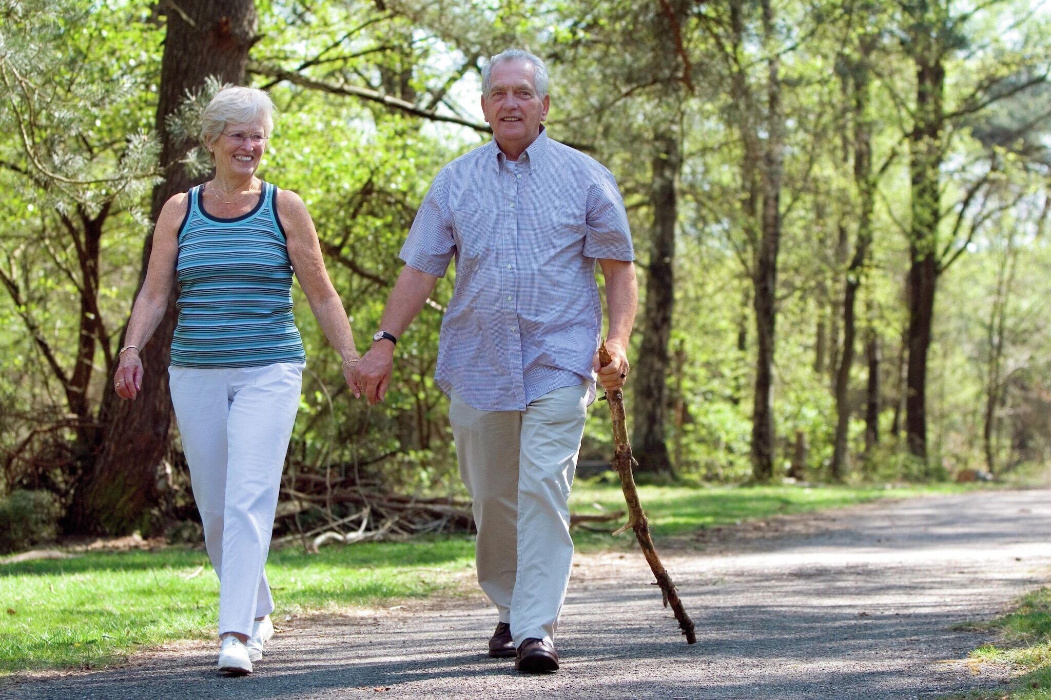 Um casal idoso caminhando por um caminho pavimentado em uma área arborizada.