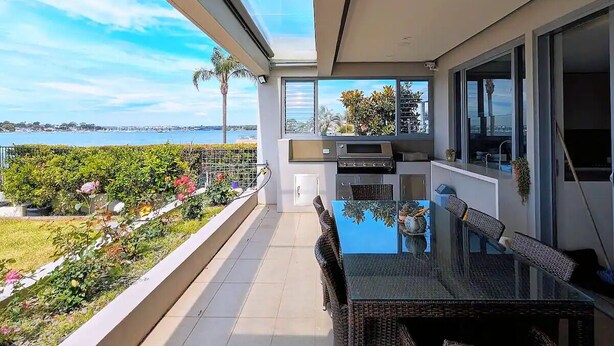 A balcony with a glass table and chairs, a grill, and a view of the sea. - Waterfront Oasis - Panoramic Views | Infinity Pool (Kogarah Bay)