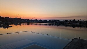 A serene infinity pool with a wooden deck, overlooking a calm body of water and a cityscape at sunset. - Waterfront Oasis - Panoramic Views | Infinity Pool (Kogarah Bay)