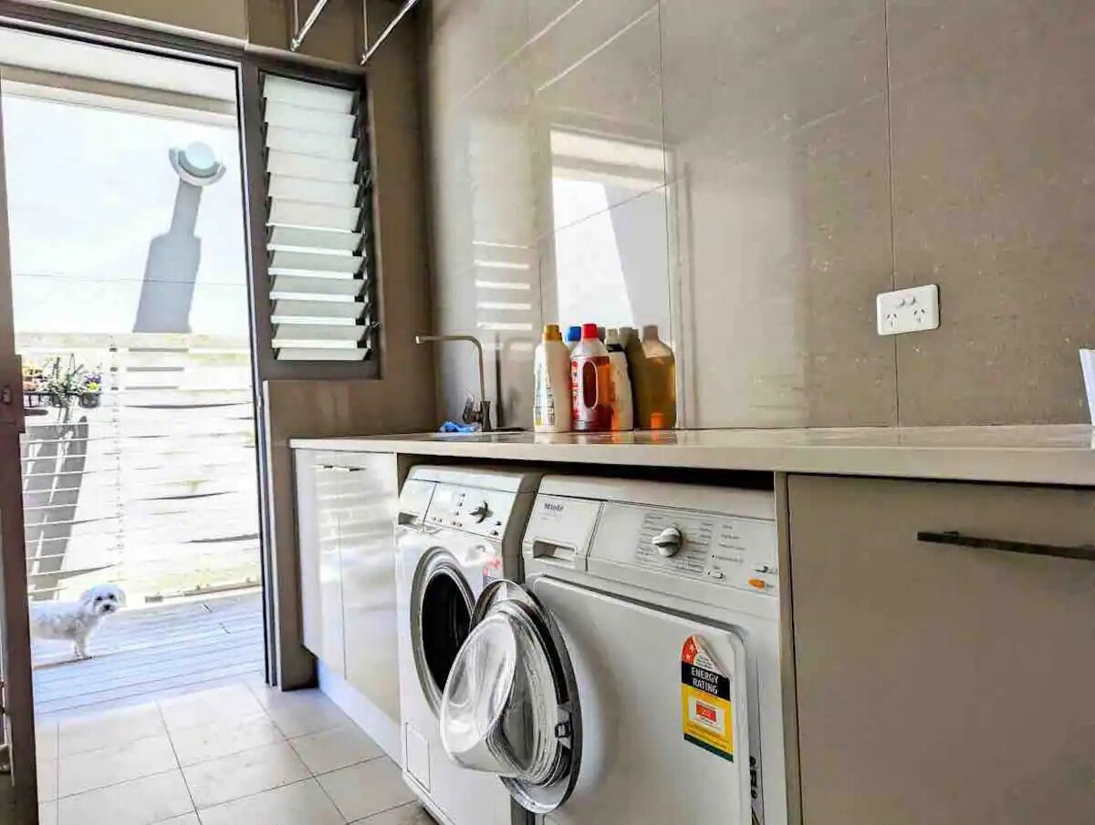 A modern laundry room with a front-loading washing machine, a drying rack, and a window with blinds.