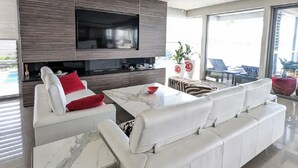 A modern living room with a large flat-screen TV, white sofas, a marble coffee table, and a dining area with a glass table. - Waterfront Oasis - Panoramic Views | Infinity Pool (Kogarah Bay)