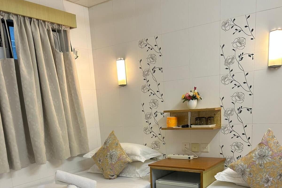 A hotel room with two beds, a small desk, a nightstand, a wall-mounted shelf with decorative items, and a window with curtains.