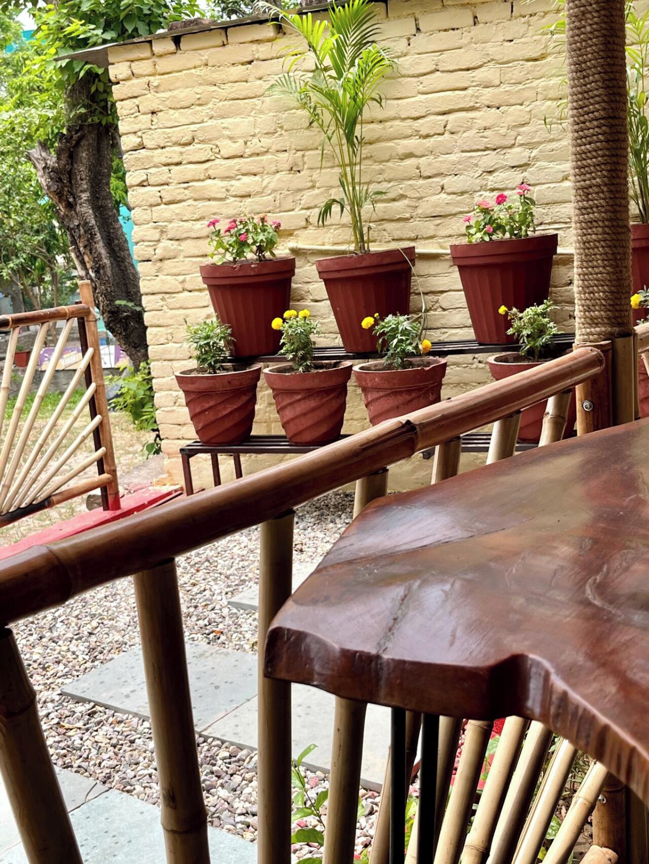 Una mesa y sillas de madera en una zona de estar al aire libre, con plantas en macetas y un muro de ladrillo al fondo.