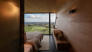 A bedroom with a large window, a bed, a bedside table, and a view of a green landscape. - The Skybarrel (Buninyong)