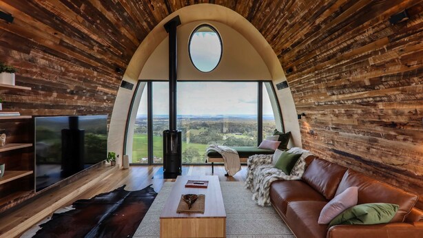 A cozy living room with a large arched window, a leather sofa, a coffee table, and a flat-screen TV. - The Skybarrel (Buninyong)