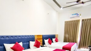Een hotelkamer met twee bedden, een blauw hoofdbord, rode en witte kussens, een rode deken, een plafondventilator en een raam met gordijnen.