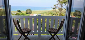 Ein Balkon mit zwei Holzstühlen und einem weißen Geländer, der auf eine Küstenlandschaft mit viel Grün und Blick aufs Meer blickt.
