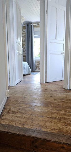 Un couloir au plancher de bois menant à une chambre dont un mur est en pierre et qui possède une fenêtre avec des rideaux.
