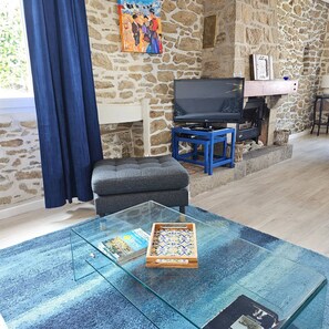 A living room with a stone wall, a television on a stand, a glass coffee table with magazines and a board game, and a blue curtain.