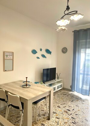 Ein Essbereich mit einem Holztisch, Stühlen, einem Fernseher auf einem weißen Schrank, Wandkunst mit Fischmotiven, einer Uhr und einem Fenster mit blauem Vorhang.