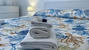 Un lit bien fait, recouvert d’une courtepointe à motifs floraux bleus et blancs, une serviette pliée et un tableau représentant un paysage marin accroché au mur.