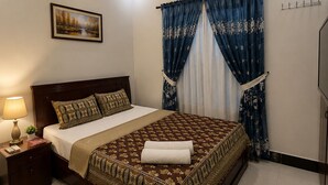 Une chambre d’hôtel comprenant un lit, une table de chevet, une lampe, un ventilateur de plafond, un appareil de climatisation, un tableau et une fenêtre avec des rideaux.