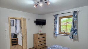 Ein Schlafzimmer mit Bett, Fernseher, einem Holzkoffer, einem Fenster mit Vorhängen und einer Tür zu einem anderen Raum.