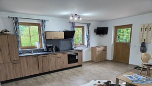 Eine moderne Küche mit Holzschränken, einem eingebauten Ofen und einer Kaffeemaschine. Zwei Fenster mit Vorhängen, ein an der Wand befestigtes Flachbildfernsehen und eine Holztür.