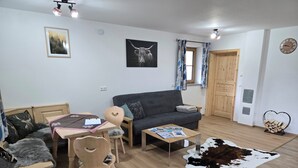 Ein gemütliches Wohnzimmer mit einer Couch, einem Esstisch mit Stühlen, einem Couchtisch mit Zeitschriften, einem Kuhfellteppich und einer Holztür.