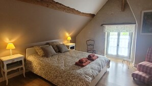 Une chambre avec un plafond en pente, un lit avec une literie à motifs, deux lampes de chevet, une chaise et une fenêtre avec un rideau.