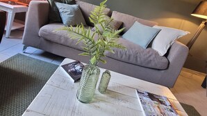 Un salon avec un canapé gris, une petite table sur laquelle repose un magazine, un vase avec une plante et un tableau accroché au mur.
