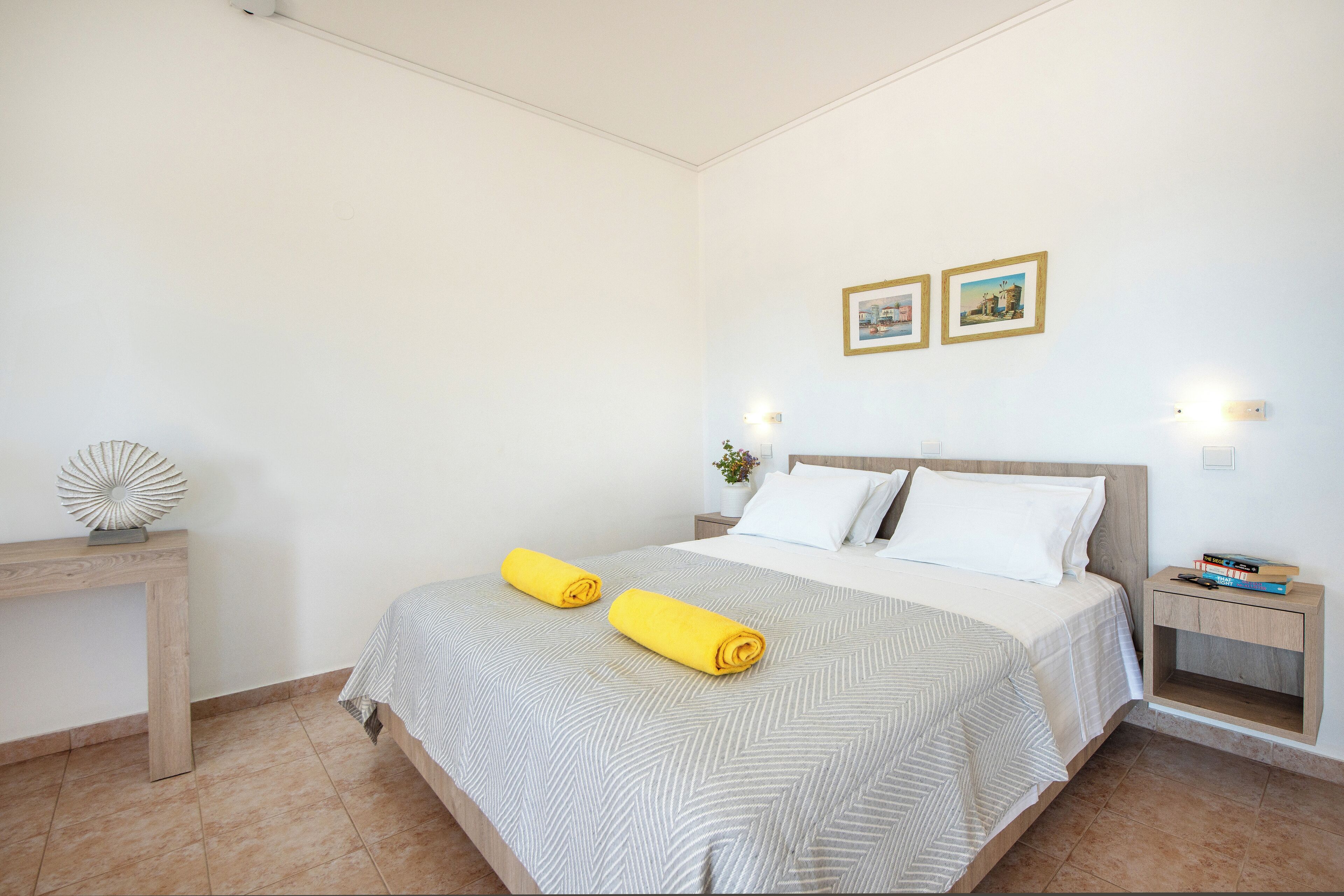 A neatly made bed with a striped bedspread and two rolled towels, a bedside table with a decorative object, and two framed pictures on the wall.