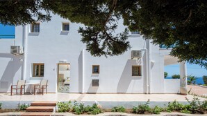 Un bâtiment blanc doté de nombreuses fenêtres, un patio avec des meubles d’extérieur et un mur de soutènement en pierre agrémenté de plantes.