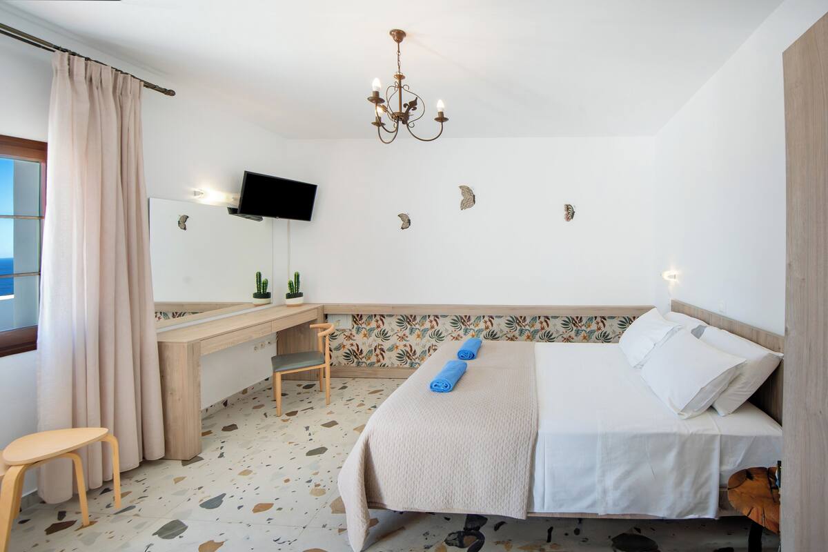 A hotel room with a bed, a desk, a chair, a TV, and a view of the sea.