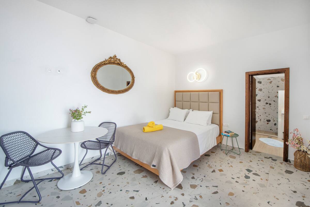 A bedroom with a bed, bedside table, chair, and a small round table with a plant.