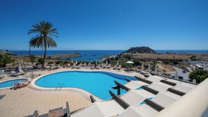 Une piscine d’hôtel avec des chaises longues et un palmier, offrant une vue sur un paysage côtier et une structure ressemblant à un fort.