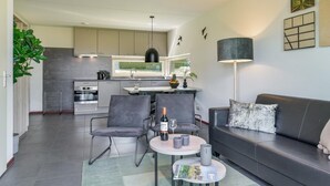A modern kitchen with grey cabinets, a dining area with grey chairs, a black sofa with a cushion, and a table with a bottle and glasses. - Reevallis Holiday House, Vijlen (Vijlen)