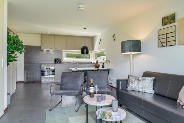 A modern kitchen with grey cabinets, a dining area with grey chairs, a black sofa with a cushion, and a table with a bottle and glasses.