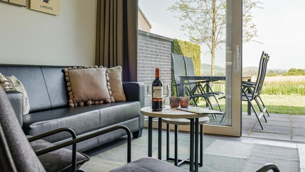 A modern living room with a black leather sofa, a small round table with a bottle of wine, and a glass. Outside, there's a glass door leading to a patio with a table and chairs. - Reevallis Holiday House, Vijlen (Vijlen)