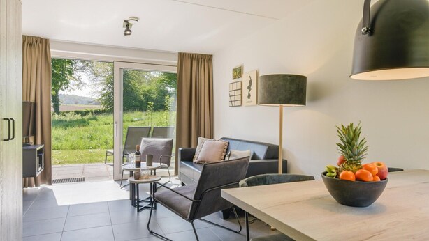 Ett modernt vardagsrum med en soffa, ett soffbord och ett matbord. Det finns ett stort fönster med gardiner, en svart lampa och en skål med frukt på bordet.